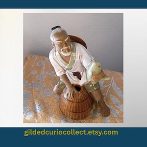 May include: A ceramic figurine of a bearded person wearing a white tunic with green trim and brown shorts, kneeling beside a brown woven-style basket while holding a small green fish. The figure has dark hair tied back and a light skin tone.