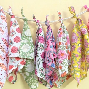 May include: A series of nine square fabric napkins hanging from a white cord, each featuring distinct block-printed patterns in vibrant colours including red, orange, green, pink, and yellow with floral, paisley, and fish motifs on white backgrounds.
