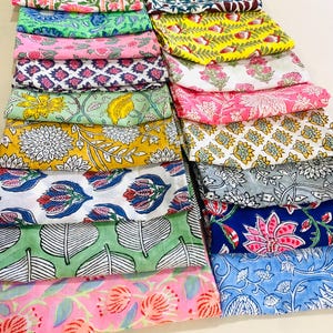 May include: A collection of folded cotton fabric squares featuring various block-printed floral and botanical patterns in vibrant colours including yellow, pink, blue, green, and grey. Each piece displays distinct motifs like leaves, petals, and stems.