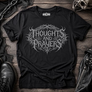 以下が含まれることがあります： 「THOUGHTS AND PRAYERS」という文字が複雑な装飾フォントで描かれたグレーのグラフィックプリントが施された黒の半袖Tシャツ。文字は棘のある蔓のような円形の枠で囲まれており、Tシャツにはクルーネックとブランドラベルが付いています。