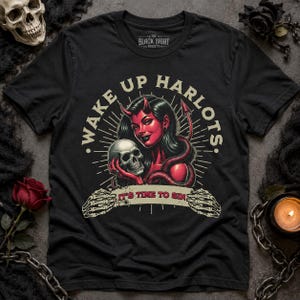 Op de afbeelding: Een zwart t-shirt met korte mouwen en een afbeelding van een roodkleurige duivelsfiguur die een menselijke schedel vasthoudt. De tekst luidt WAKE UP HARLOTS en IT'S TIME TO SIN. Het ontwerp bevat een skelet-handbanner en stralende lijnen.