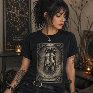 May include: A black short-sleeved t-shirt featuring a rectangular graphic print of a tarot card design. The print depicts two identical figures standing back-to-back, surrounded by thorny vines and stars, with the word GEMINI printed at the bottom.