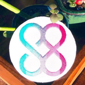 Peut inclure: Un écusson brodé rond blanc avec un symbole infini stylisé dans un dégradé de turquoise, violet et rose. Le patch est placé près d'une petite plante en pot et d'une surface en bois.