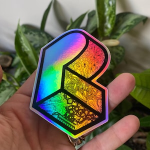 May include: A holographic sticker with a black outline and a colorful, intricate design. The sticker features a stylized letter "R" with a black background and a rainbow iridescent finish.