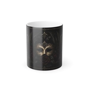 May include: A black ceramic mug featuring a gold and cream-colored masquerade mask design. The mask is surrounded by dark red roses and feathers. The mug has a white interior and a black exterior with a decorative border.
