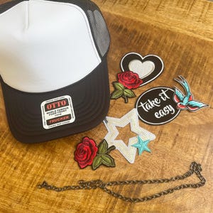 May include: A black and white trucker hat with an Otto brand sticker on the brim, accompanied by several embroidered patches including a red rose, a heart, a star, a swallow, and a circular patch with the text take it easy, plus a metal chain.