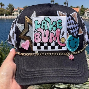 May include: A black trucker hat featuring a central white patch with the text LAKE BUM in teal and pink letters. The hat is decorated with a gold chain, a pink heart, a teal smiley face, a checkered flag, and a large gold sequined star.