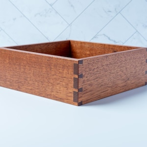 May include: A square wooden box with a medium brown finish and visible dovetail joinery at the corners. The box features four straight sides and an open top, showcasing the natural grain of the wood and the contrasting darker wood used for the joints.
