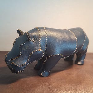 Op de afbeelding: Een donkerblauw lederen nijlpaardfiguur opgebouwd uit meerdere gestikte panelen. Het dierenfiguur is voorzien van opvallende lichtgekleurde draaddetails langs de naden, die de vorm van de kop, het lichaam en de poten in een gesegmenteerd ontwerp definiëren.