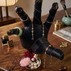 May include: A black, hand-shaped jewelry display stand featuring visible tan stitching along the edges. The stand is adorned with a light green jade-style ring, two delicate gold-toned chain bracelets, and a silver-toned ring on the fingers.