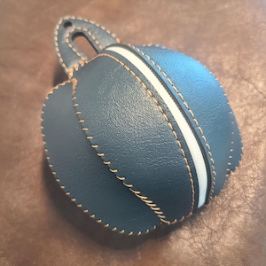 May include: A round, dark blue pouch with a textured surface and a white zipper closure. The pouch features prominent tan stitching along the seams and includes a small, integrated loop at the top for hanging or attachment purposes.