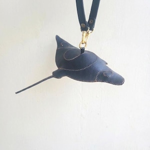 May include: A black leather stingray-shaped keychain featuring visible light-colored stitching along the edges and body. The stingray is attached to a black leather strap by a gold-toned metal swivel lobster clasp and a small metal ring.