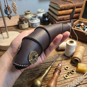 May include: A dark brown textured leather case shaped like a fish, featuring a wrap-around leather cord and a circular brass coin with Chinese characters attached to the side. The case is stitched with white thread along the edges.