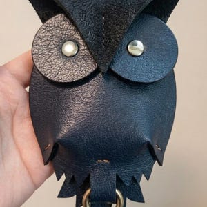 Peut inclure: Un accessoire en cuir noir en forme de hibou doté de deux patchs oculaires circulaires texturés avec des rivets métalliques argentés, un corps en cuir lisse et un anneau métallique doré fixé au centre inférieur avec des coutures visibles.