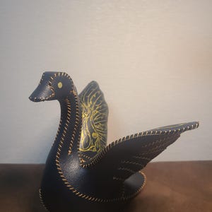 May include: A black leather swan figurine featuring prominent tan stitching along the edges of its body, neck, and wings. The wings display an intricate gold-coloured leaf or feather pattern printed on the surface, and the eye is a small gold dot.