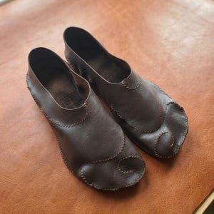 Op de afbeelding: Een paar donkerbruine leren schoenen met een gespleten neus en zichtbare stiksels langs de randen. De schoenen hebben een laag profiel en een gestructureerd oppervlak met een zichtbare merkstempel op de binnenzool.