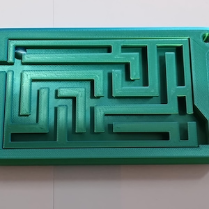 Puede incluir: Un rompecabezas de laberinto rectangular de plástico impreso en 3D en color verde azulado, con una red compleja de paredes y caminos elevados. Una pequeña bola de metal plateada se encuentra dentro de las pistas. El marco tiene un borde liso.