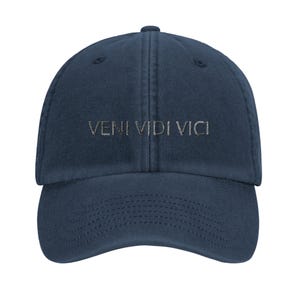 May include: A navy blue baseball cap featuring the text VENI VIDI VICI embroidered in a light grey thread across the front panel. The cap has a structured crown, a curved brim with visible stitching, and a button at the top centre.