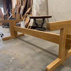 May include: A light brown wooden table base featuring a long horizontal support beam connected to two vertical end supports with wide, flat feet. The structure is constructed from thick, natural-toned timber planks with visible wood grain patterns.