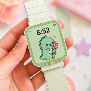 Puede incluir: Un smartwatch de papel verde claro con un dinosaurio de dibujos animados sosteniendo una flor en la pantalla. Se muestra la hora 6:52. La correa del reloj es verde claro con un patrón de hojas.