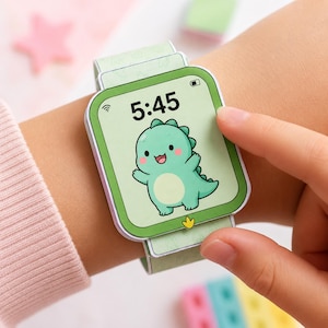 Könnte beinhalten: Eine hellgrüne Papier-Smartwatch mit einem Cartoon-Dinosaurier auf dem Bildschirm. Die angezeigte Uhrzeit ist 5:45 Uhr. Die Uhr befindet sich an einem Handgelenk, und ein Finger berührt den Bildschirm.