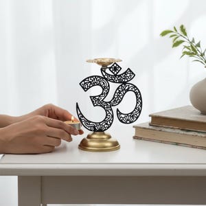 Könnte beinhalten: Ein schwarzer Metall-Kerzenhalter in Form des Om-Symbols mit einem goldfarbenen Fuß und einer kleinen runden Platte für ein Teelicht. Das Om-Design weist filigrane florale Ausschnitte auf und steht auf einem glatten, goldenen Sockel.