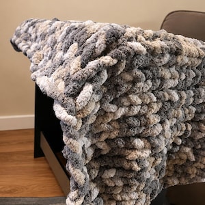 May include: A chunky knit blanket in shades of gray, white, and tan is draped over a dark surface. The thick, textured yarn creates a cozy, inviting look. This soft blanket is perfect for adding warmth and style to any living space.
