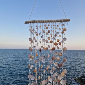 Puede incluir: Una decoración de pared que presenta un trozo de madera flotante horizontal con múltiples cuerdas blancas verticales suspendidas. Cada cuerda está adornada con pequeñas cuentas blancas y varias conchas naturales en tonos bronce, marrón y blanco.