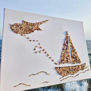 Può includere: Una tela bianca con un mosaico di piccole conchiglie marroni e beige che formano la sagoma di una barca a vela e dell'isola di Cipro, con una scia di conchiglie che collega le due figure e sottili linee di conchiglie che raffigurano onde.