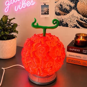 Op de afbeelding: Een bolvormige lamp met een feloranje, getextureerd oppervlak met wervelende vlamachtige patronen en een opvallende groene steel aan de bovenkant. De lamp staat op een ronde grijze voet en wordt van binnenuit verlicht met een warme gloed.