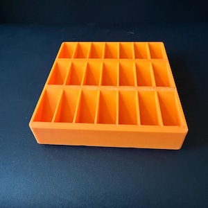 May include: A bright orange rectangular storage tray featuring a grid of eighteen individual compartments arranged in two rows of nine. The organizer has a solid base and vertical dividers that create uniform slots for small items.