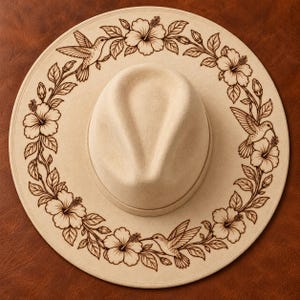 Puede incluir: Un sombrero beige de ala ancha con un diseño grabado con láser en color marrón oscuro en el ala. El patrón consiste en una disposición circular continua de flores de hibisco, hojas y tres colibríes en vuelo alrededor de la copa.