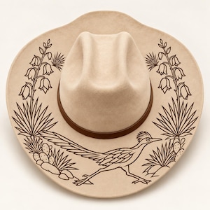 May include: A beige felt cowboy hat featuring a dark brown band around the crown. The wide brim is decorated with a dark brown line art illustration of a roadrunner bird, desert cacti, and yucca plants arranged in a symmetrical pattern.