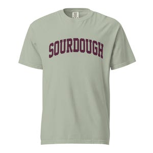 May include: A sage green short-sleeve t-shirt featuring the word SOURDOUGH printed in a dark maroon, arched collegiate-style font across the chest. The garment has a crew neckline and a standard fit, with a visible brand label at the inner collar.
