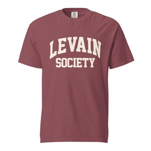 May include: A short-sleeve crew neck t-shirt in a muted dark red color. The front of the shirt features the text "LEVAIN SOCIETY" printed in a large, cream-colored, arched collegiate-style font across the center of the chest area.