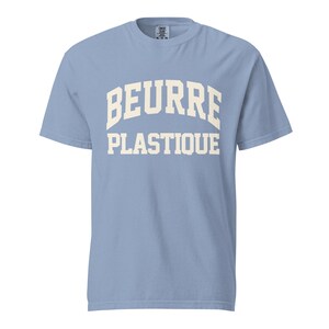 May include: A light blue short-sleeve t-shirt featuring the text BEURRE PLASTIQUE printed in a large, cream-colored, arched collegiate-style font across the chest. The garment has a crew neckline and a standard fit, with a visible brand label.