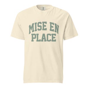 May include: A cream-colored short-sleeve t-shirt featuring the text MISE EN PLACE printed in a sage green collegiate-style font across the chest. The garment has a crew neckline and a standard fit, with a visible brand label at the inner collar.