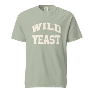 May include: A sage green short-sleeved t-shirt featuring the text WILD YEAST printed in a large, cream-colored, arched collegiate-style font across the chest. The garment has a crew neckline and a standard fit, showcasing a solid color design.