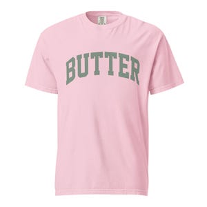 May include: A light pink short-sleeve t-shirt featuring the word BUTTER printed in a large, arched, sage green collegiate-style font across the chest. The garment has a crew neckline and a standard fit, with a visible brand label at the neck.