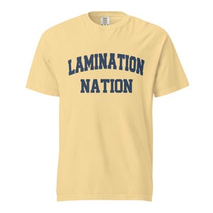 May include: A pale yellow short-sleeve t-shirt featuring the text LAMINATION NATION printed in dark blue block letters across the chest. The top line of text is arched, while the bottom line is straight, centered on the garment.