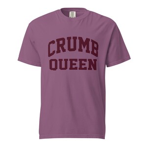 May include: A mauve short-sleeved t-shirt featuring the text CRUMB QUEEN printed in dark maroon block lettering across the chest. The shirt has a crew neckline and a standard fit, with the brand label visible at the inside of the collar.