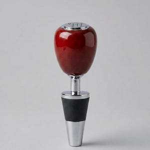 May include: A wine bottle stopper with a polished, deep red, oval-shaped top. The stopper has a silver-coloured metal stem and base, and a black rubber seal. The top has a silver-coloured, circular design.
