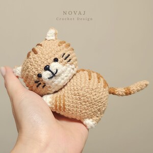 May include: A small, handmade crochet cat toy featuring light tan yarn with darker brown striped markings, a white muzzle, and black embroidered eyes and mouth. The text "NOVAJ Crochet Design" is visible at the top of the image.