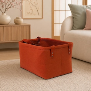 May include: A rectangular storage basket in a vibrant burnt orange colour, constructed from a felt-like fabric. The basket features two matching orange handles securely attached to the sides with silver-toned metal rivets at each connection point.