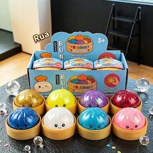 May include: A display box of Rainbow Mystery squishy bun toys with eight individual colorful, glittery, round squishy buns in small tan containers. The box features cartoon bun illustrations, the text "Squishy Bun," and "Will You Find The Super Rare!"