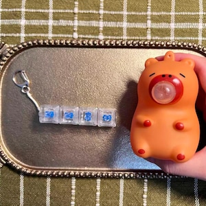 May include: A keychain featuring four clear mechanical keyboard switches with blue internal components, alongside an orange, rounded, bear-shaped squeeze toy with a red mouth area and a small, clear bubble protruding from its center.