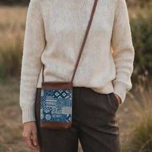 May include: A crossbody bag featuring a brown leather top and bottom with a central panel of blue and white patchwork fabric. The fabric displays various geometric patterns, including stripes, dots, and floral motifs, attached to a thin brown leather strap.