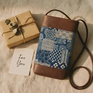 Peut inclure: Un sac bandoulière en cuir marron avec un panneau central en tissu patchwork bleu et blanc aux motifs géométriques et floraux variés. À côté du sac se trouve un coffret cadeau marron noué avec de la ficelle et une carte blanche avec le texte For You.