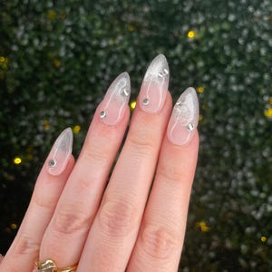 May include: Almond-shaped press-on nails featuring a clear base with a silver glitter gradient at the tips. Each nail is adorned with small, round, clear rhinestones placed near the cuticle and along the sides. A gold ring is worn on the finger.