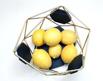 May include: A geometric gold-colored metal fruit bowl with black accents, filled with several ripe yellow lemons. The bowl has a modern design and sits on a white surface, showcasing the fresh produce.